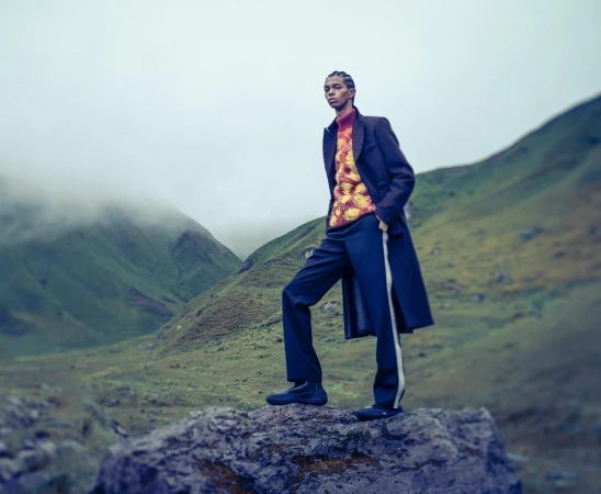 A single adult stands on a rock in outdoor fashion editorial in mountains; fog, green slopes, sweater, wool coat, pants, and shoes visible.