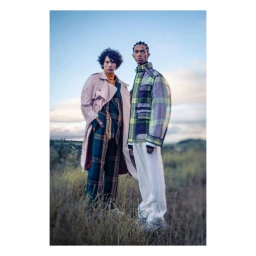 Two male models posing in a grassy field outdoors wearing plaid trench coats and trousers under a pale sky.