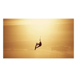 Silhouette of a kiteboarder suspended in mid-air against a bright golden sunset sky and horizon.