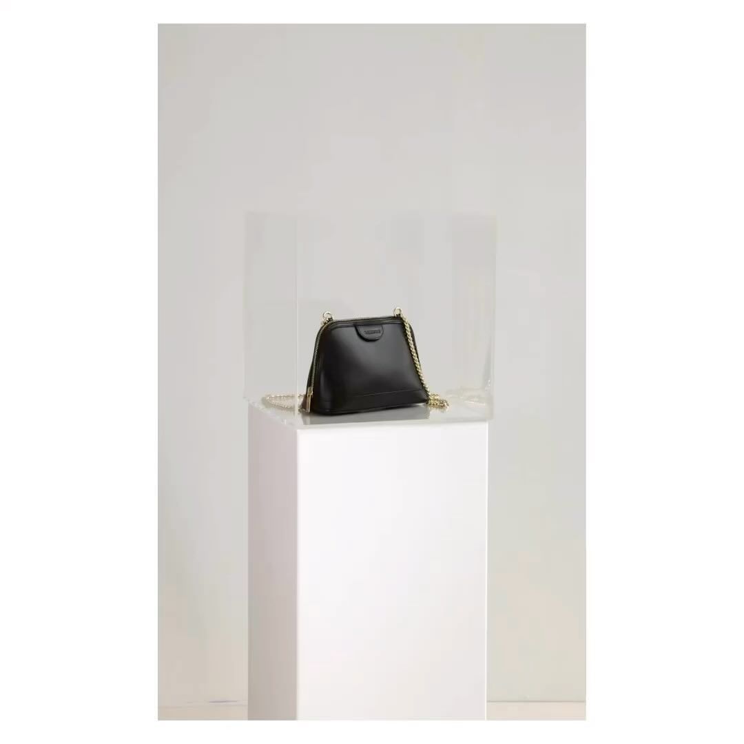 Studio shot of a black leather crossbody bag with a gold chain strap on a white platform against a gradient white background.