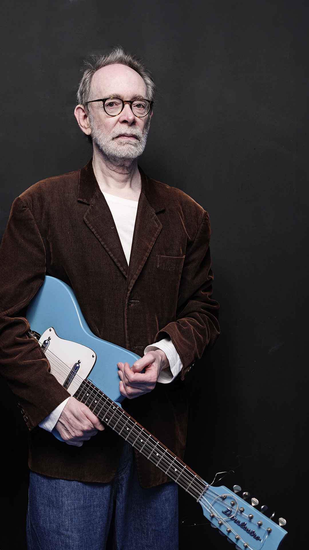 Older man with glasses stands indoors against dark backdrop holding a blue twelve string electric guitar with a corduroy jacket.