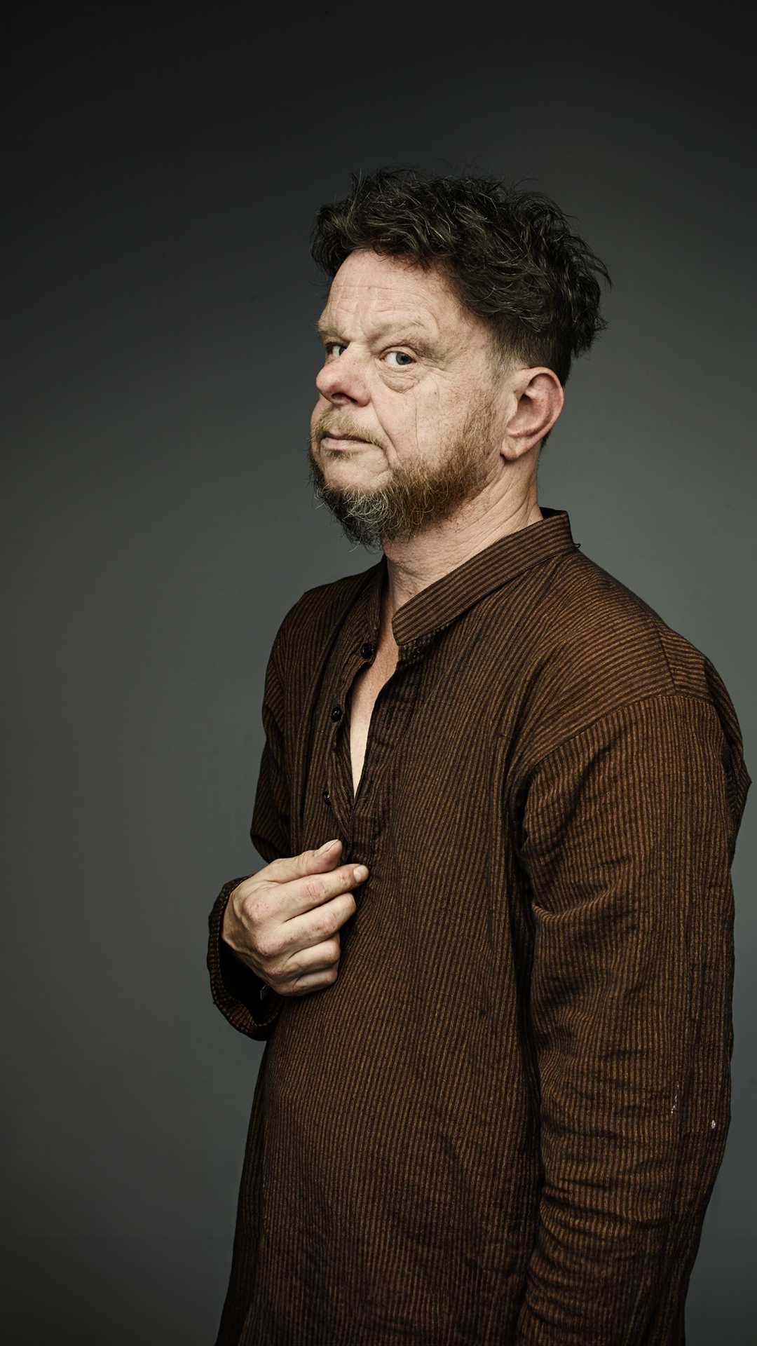 Adult male in a brown collared shirt with short hair and a beard, posing in a studio with a dark background.