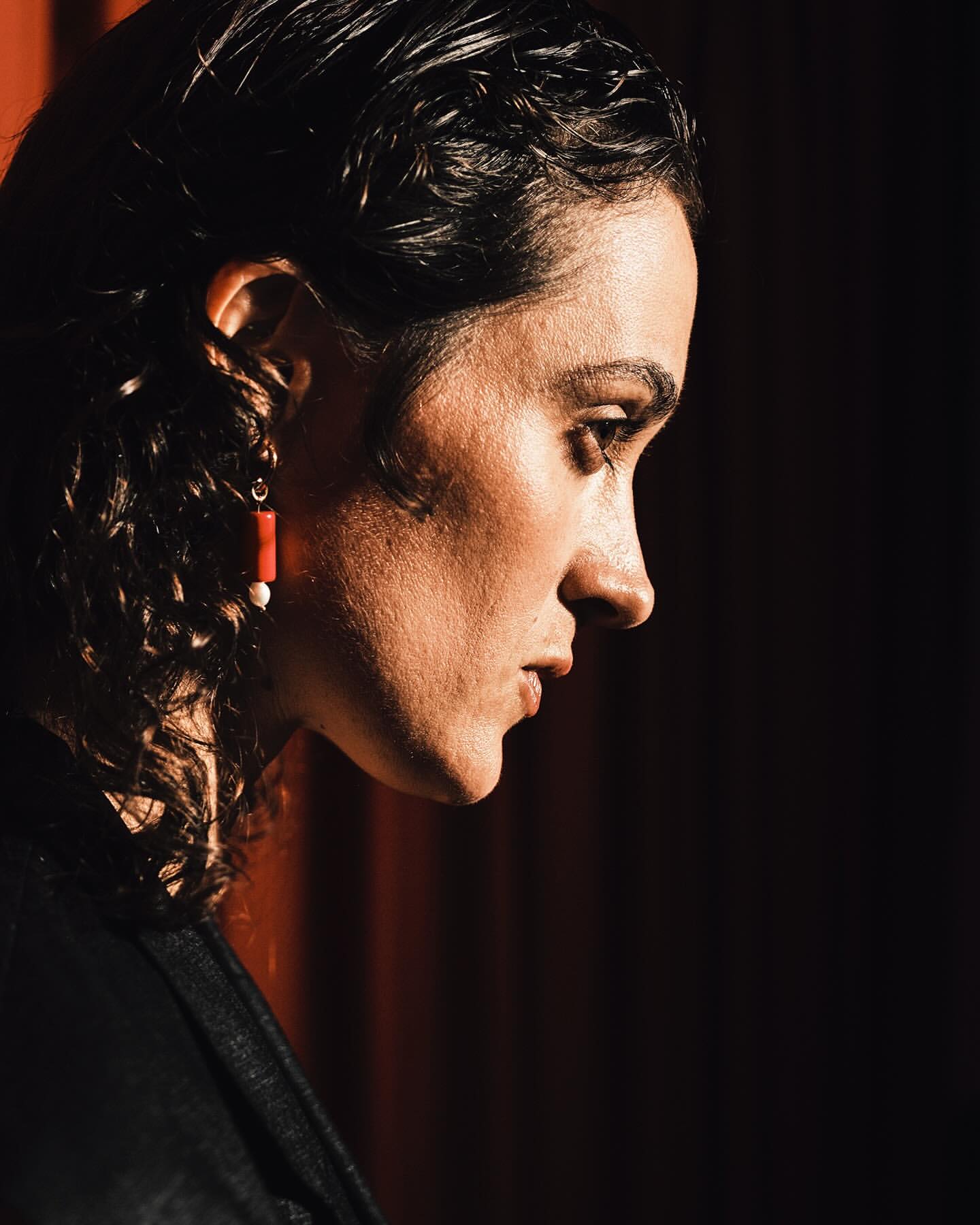 Close-up profile of adult female with dark, wet curly hair, wearing an earring, and dressed in a black top in front of a red curtain.