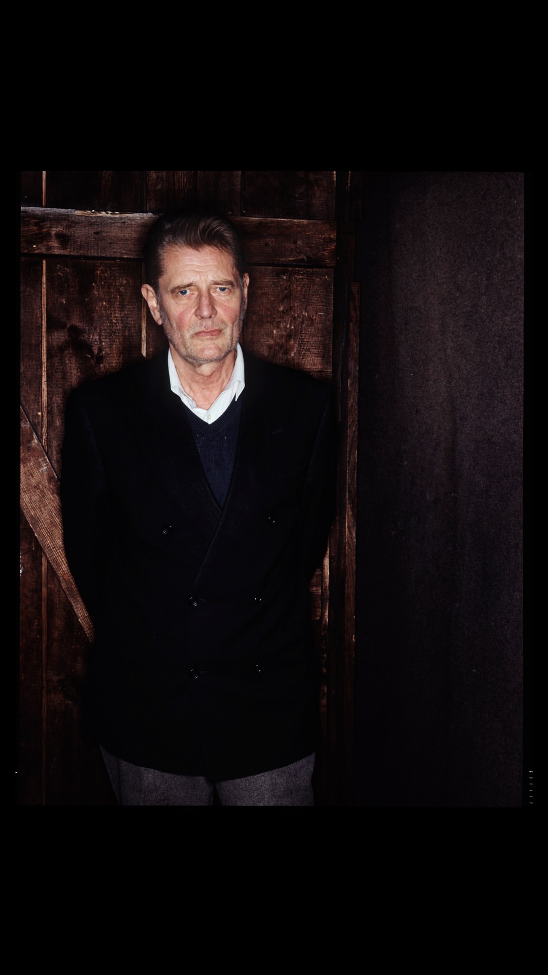 Studio portrait of a mature adult male in a dark suit standing in front of a weathered wooden door with a dark background.