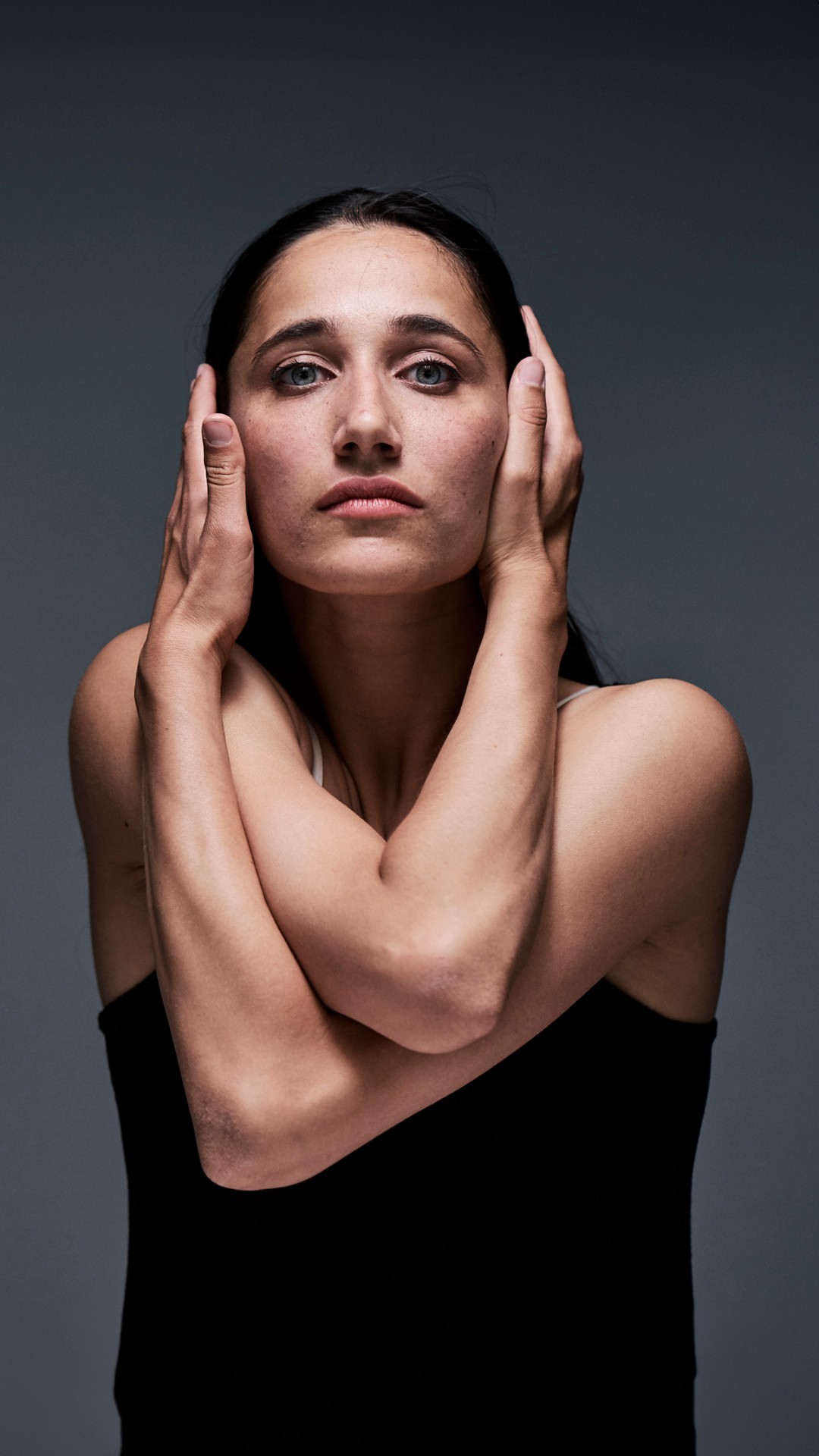 Close-up studio portrait of a woman with dark hair wearing a black top, her hands raised near her face against a grey background.