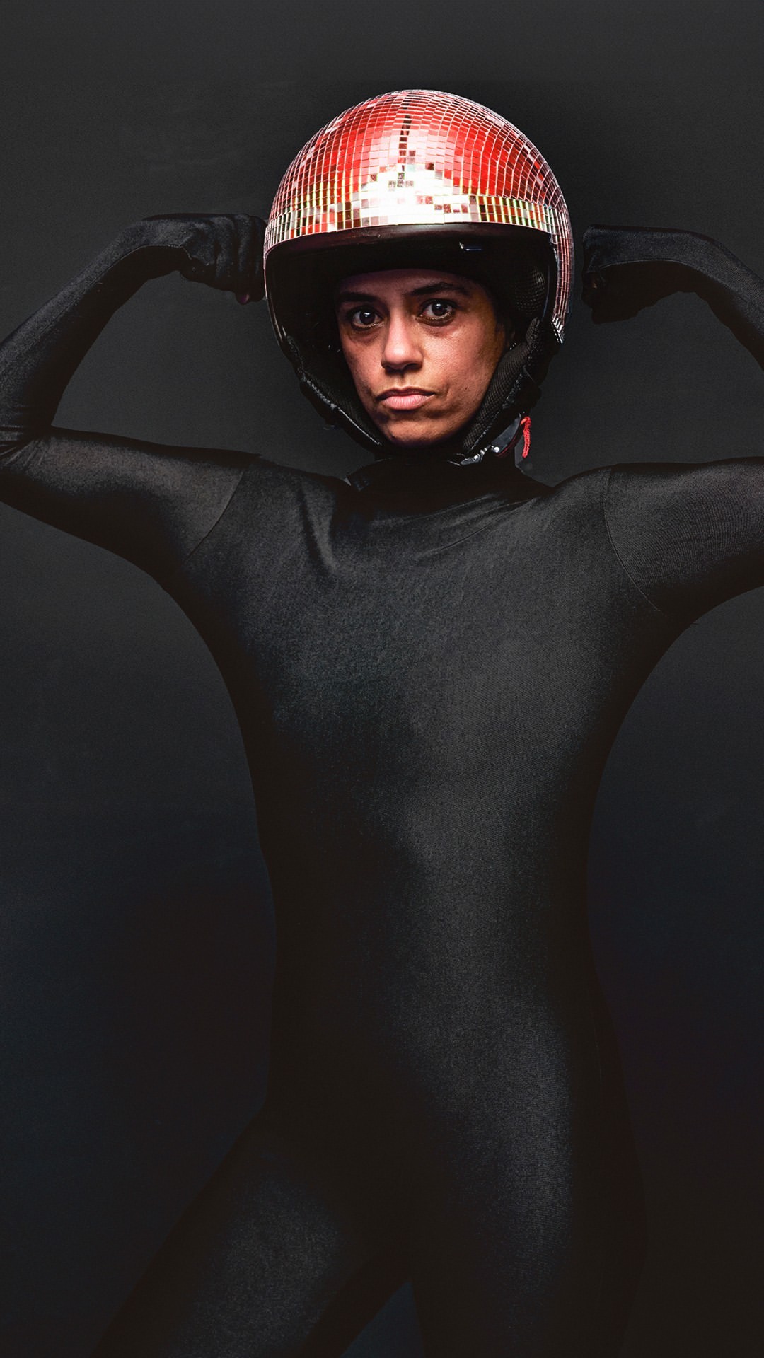 A woman in a black bodysuit and red disco ball helmet poses against a dark background in a full shot, hands raised near her head.