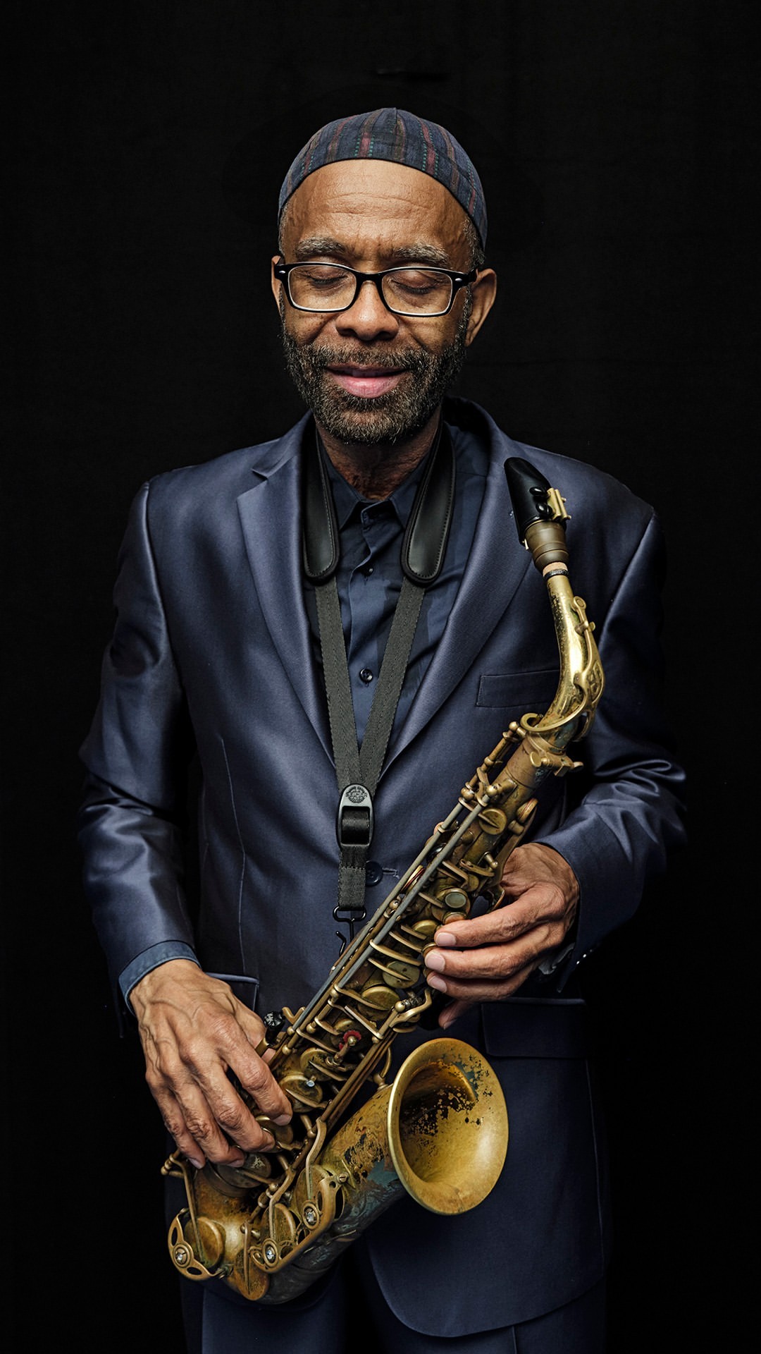 Portrait of a black man playing a saxophone in a photo studio. The musician is wearing a suit, and the background is a solid black.