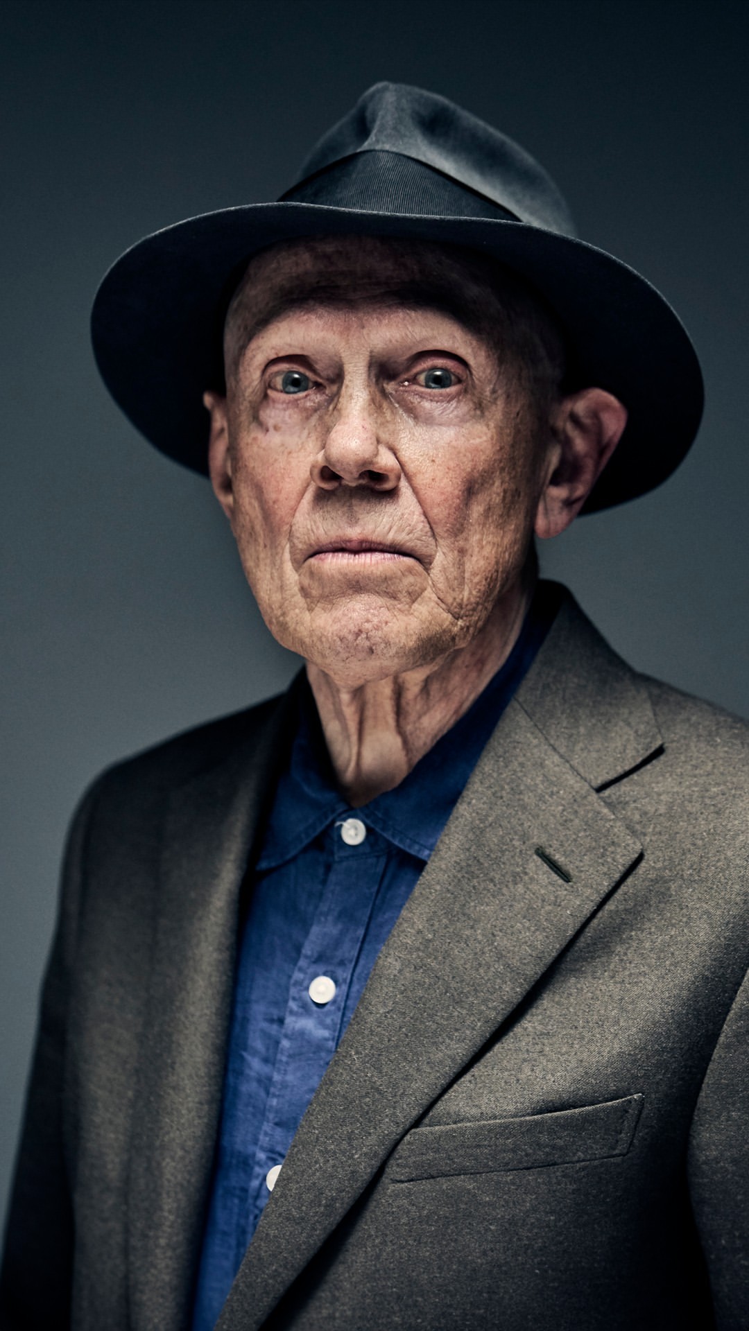 Elderly man in a fedora and tweed jacket looking at the camera against a grey backdrop in a studio portrait.