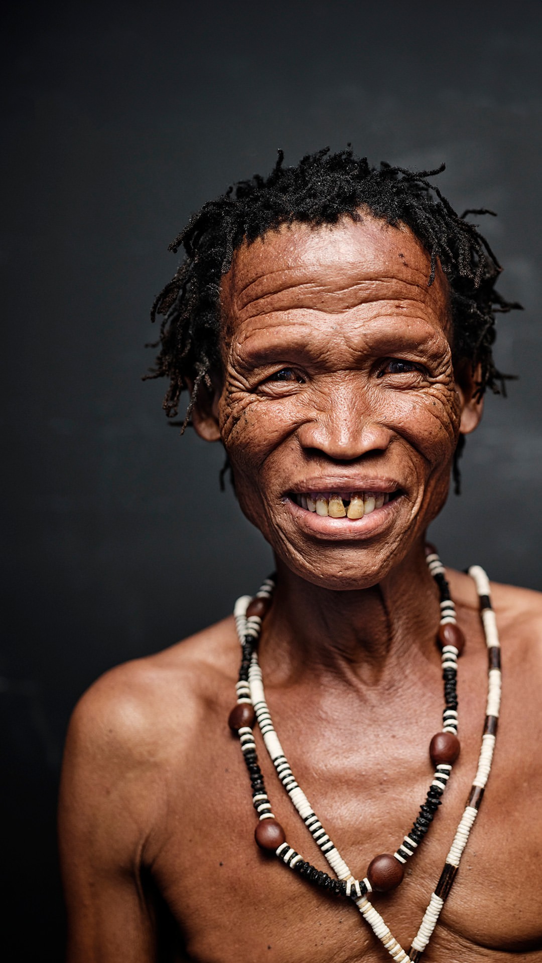 Close-up portrait of a smiling elderly African man with dreadlocks and beaded necklaces against a dark background.