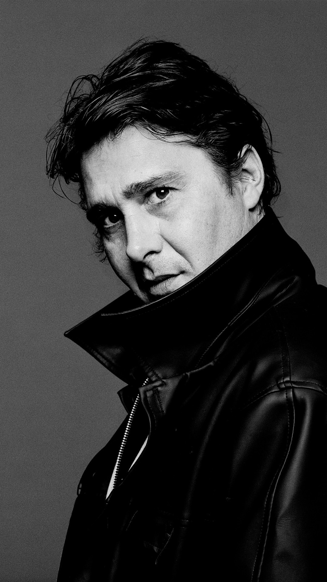 Portrait of a dark-haired man in a black leather jacket, against a grey backdrop, shot in black and white, offering a classic, stylish look.