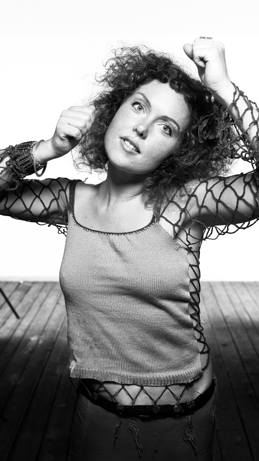Black and white portrait of a curly-haired woman in a net top, smiling with arms raised in a brightly lit indoor setting.