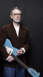 Elderly man wearing round glasses and brown blazer holding a light blue electric guitar against dark seamless background