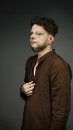 Middle-aged man with dark curly hair wearing a striped brown button-down shirt against a grey backdrop