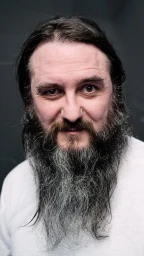 Close-up portrait of a man with a long grey beard and dark hair wearing a white t-shirt against a dark textured background.