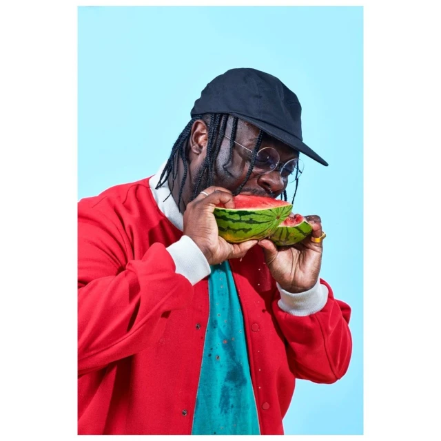 Adult male eating watermelon in a close-up studio shot. The man wears a black hat, glasses, and a red jacket against a blue background.