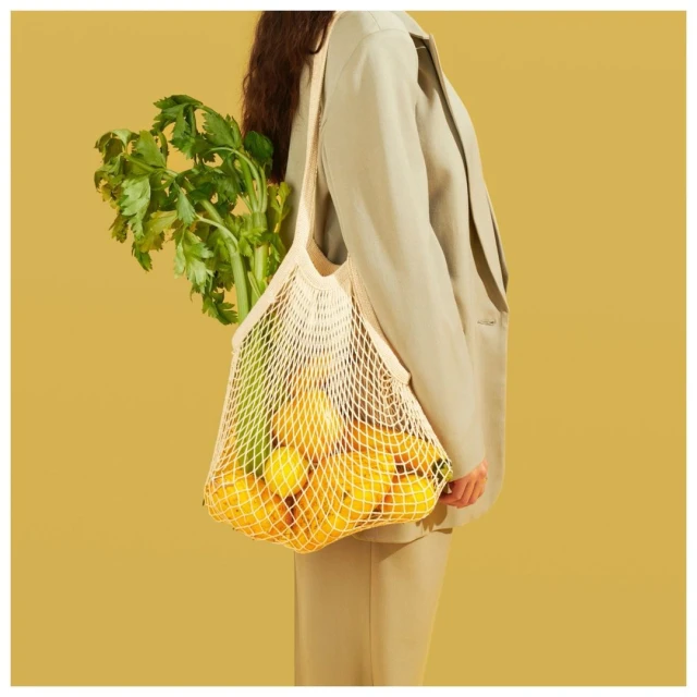 A person wearing a light brown jacket is holding a string bag filled with lemons and green leafy vegetables against a yellow background.