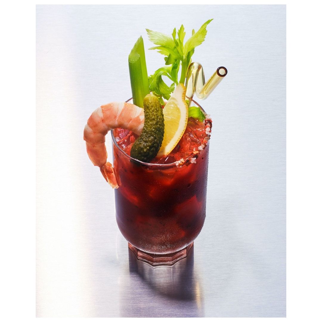 Bloody Mary cocktail in a glass with a shrimp, pickle, lemon slice, celery, and straw on a reflective metal surface.