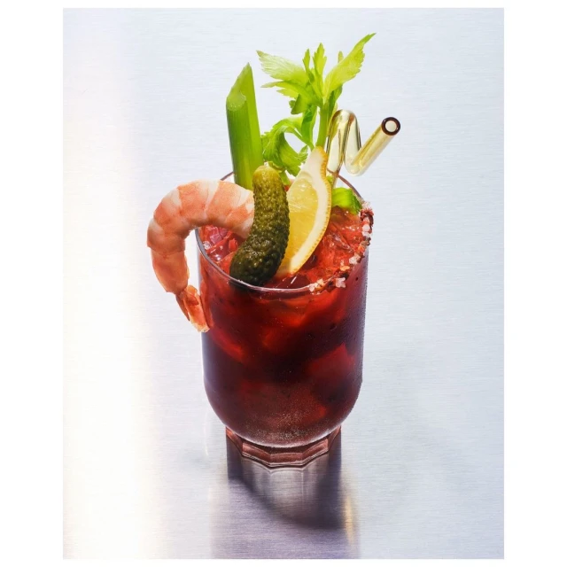 Bloody Mary cocktail in a glass with a shrimp, pickle, lemon slice, celery, and straw on a reflective metal surface.