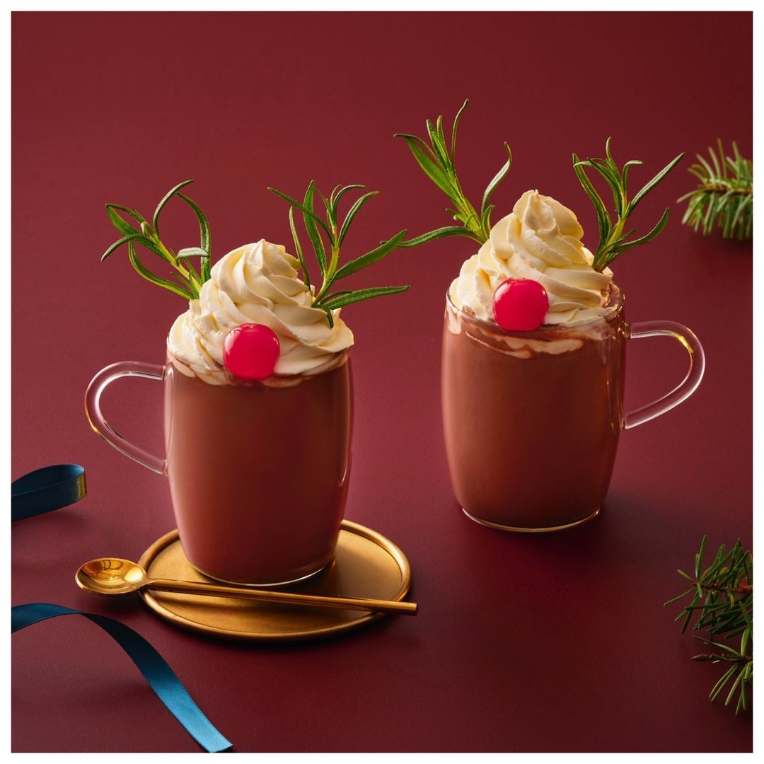 Two mugs of hot chocolate with whipped cream, a cherry, and rosemary sprigs resembling reindeer antlers against a burgundy background.