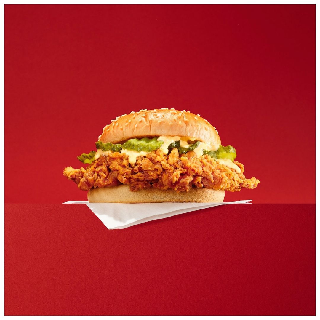 Crispy chicken sandwich with pickles and sesame seed bun against a red background, presented on a white napkin, studio shot. Food photography.