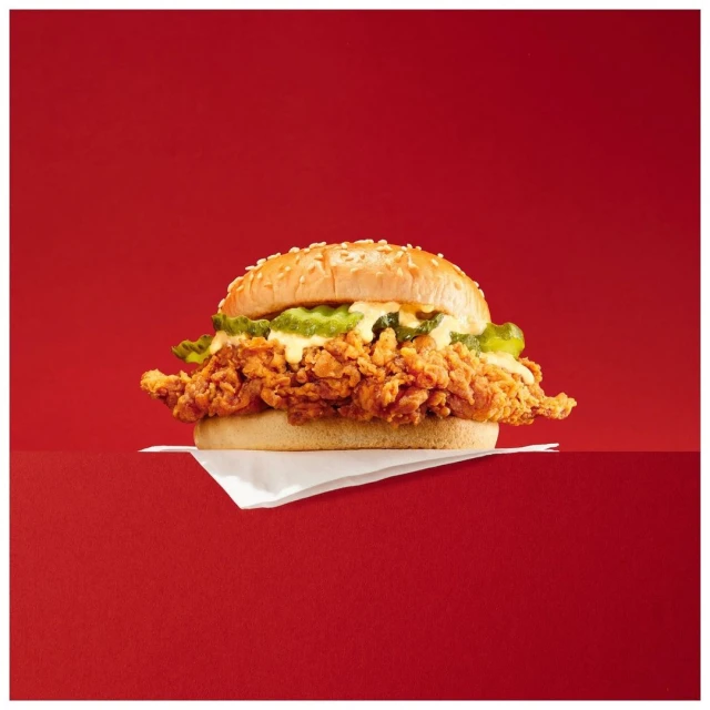 Crispy chicken sandwich with pickles and sesame seed bun against a red background, presented on a white napkin, studio shot. Food photography.