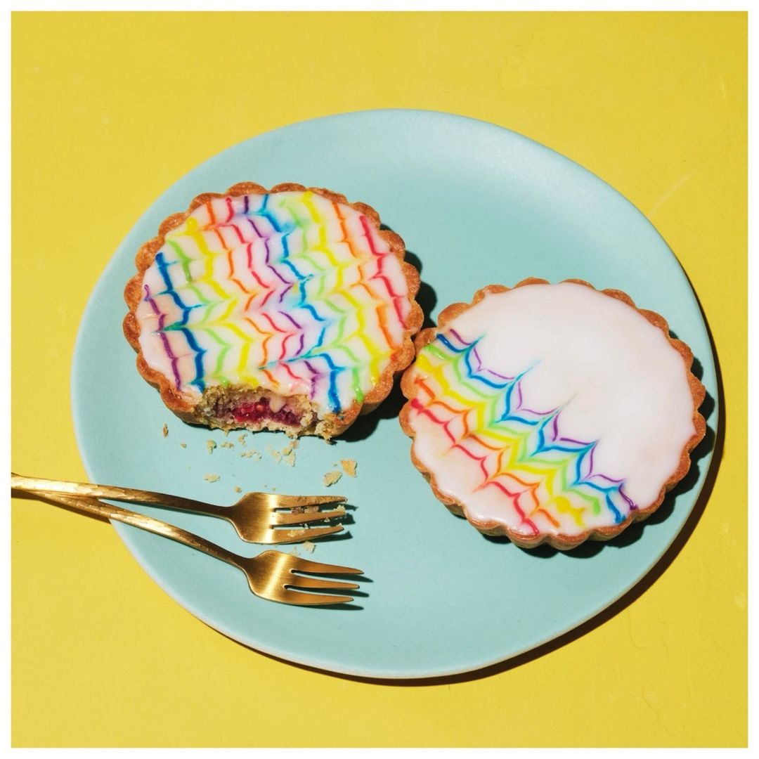 Overhead shot of two colorful tarts on a light blue plate with gold forks against a yellow background in a brightly lit setting.