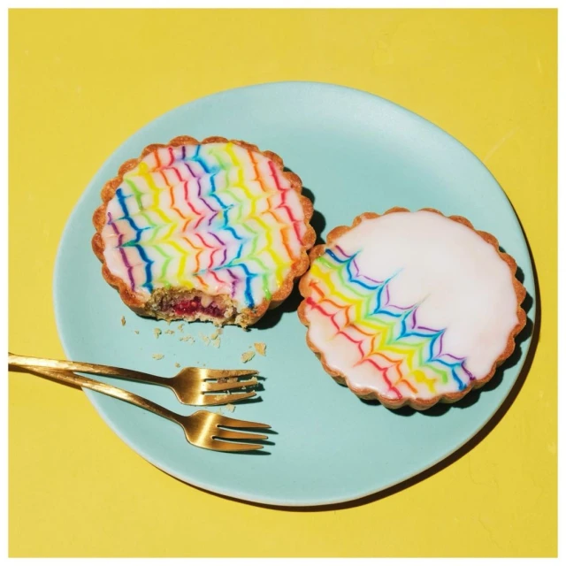 Overhead shot of two colorful tarts on a light blue plate with gold forks against a yellow background in a brightly lit setting.