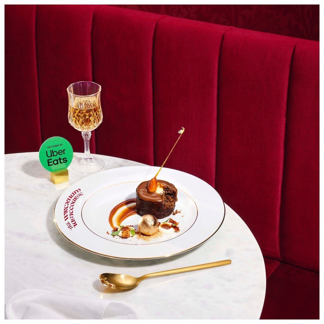 Elegant still life shot of a chocolate dessert cake on a plate with a glass of white wine, golden spoon, and Uber Eats signage.