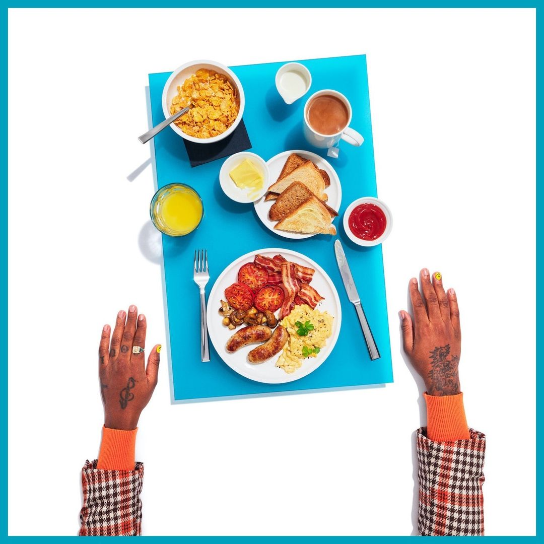 Overhead shot of a breakfast spread on a blue surface with human hands at the bottom and various breakfast foods on white plates.