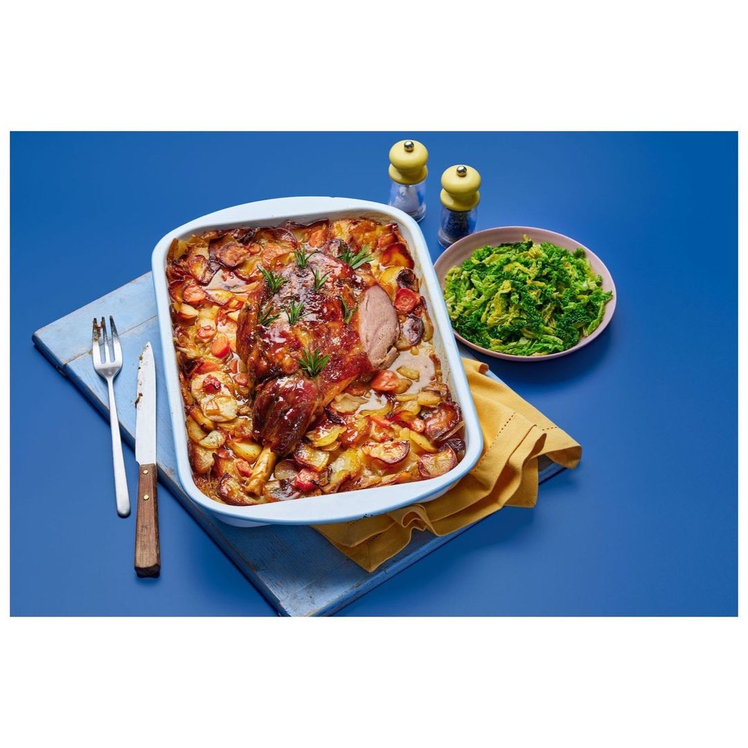 Roasted lamb and potatoes in a baking dish with a side of green vegetables on a blue background, ready to serve.
