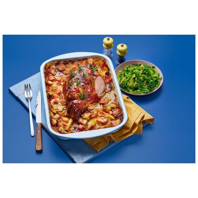 Roasted lamb and potatoes in a baking dish with a side of green vegetables on a blue background, ready to serve.
