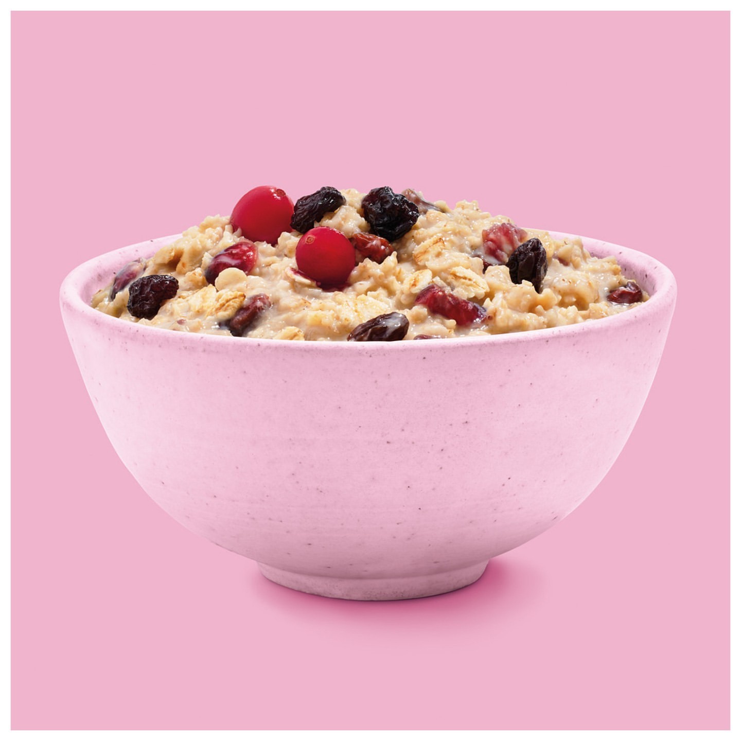 A pink ceramic bowl filled with oatmeal, red cranberries, and raisins is centered on a pastel pink background, offering a simple and appetizing view.