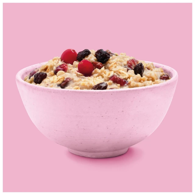 A pink ceramic bowl filled with oatmeal, red cranberries, and raisins is centered on a pastel pink background, offering a simple and appetizing view.