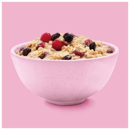 A pink ceramic bowl filled with oatmeal, red cranberries, and raisins is centered on a pastel pink background, offering a simple and appetizing view.