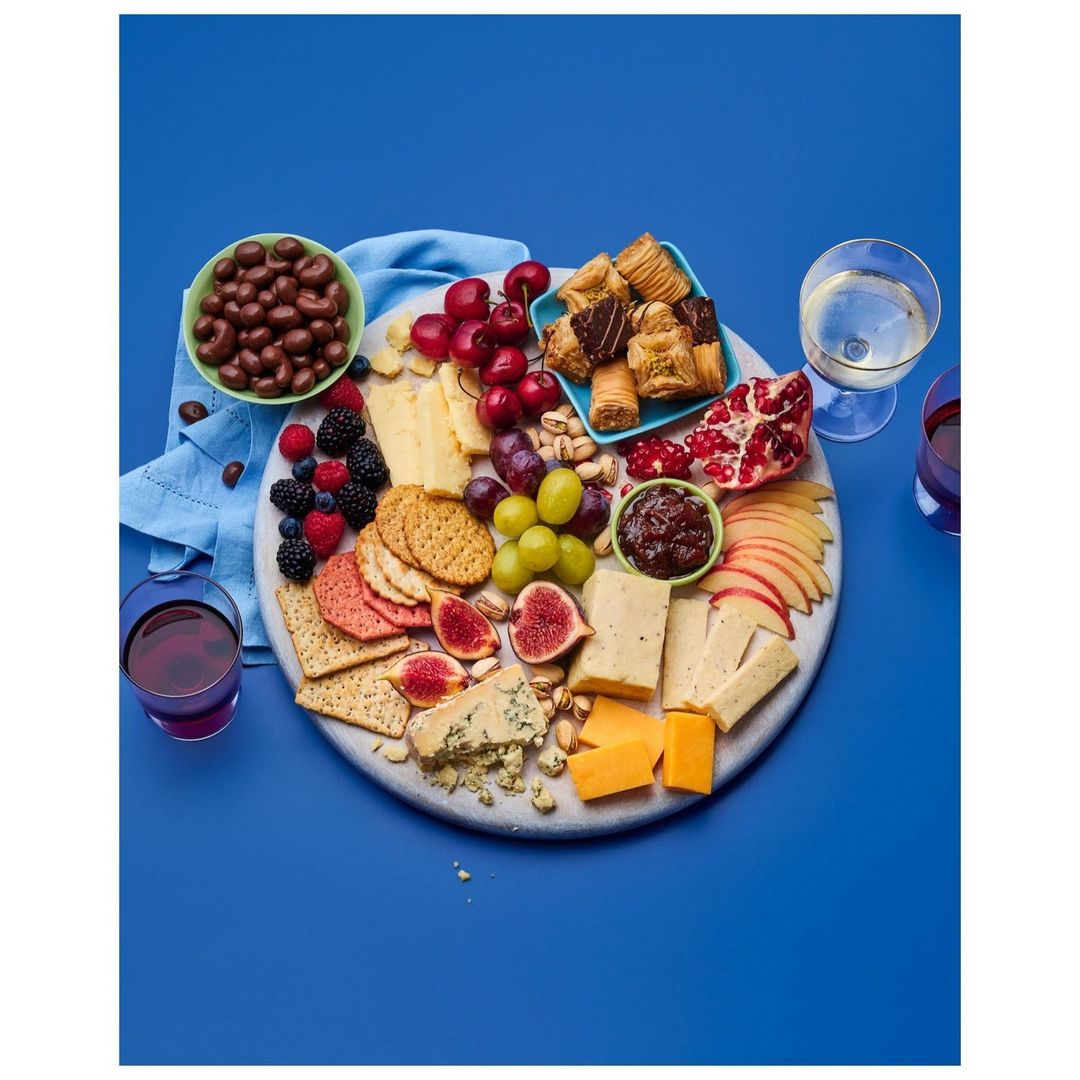 Overhead shot of a charcuterie board featuring cheese, fruit, crackers, pastries, and nuts, accompanied by glasses of red and white wine.