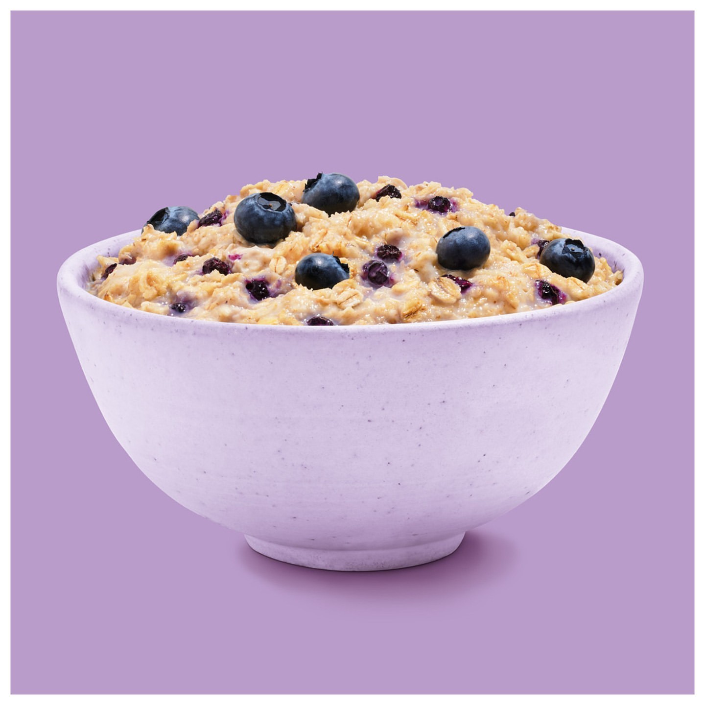 Close-up of a ceramic bowl filled with oatmeal and fresh blueberries against a solid purple background in a studio setting.