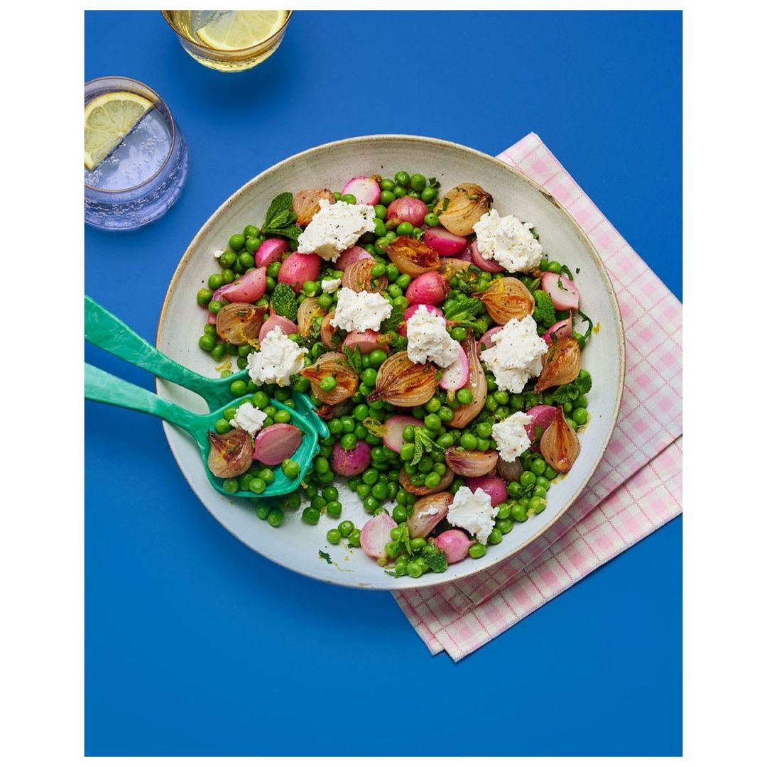 Overhead shot of a salad with peas, radishes, grilled onions, and ricotta on a plate with serving spoons, set against a blue background.