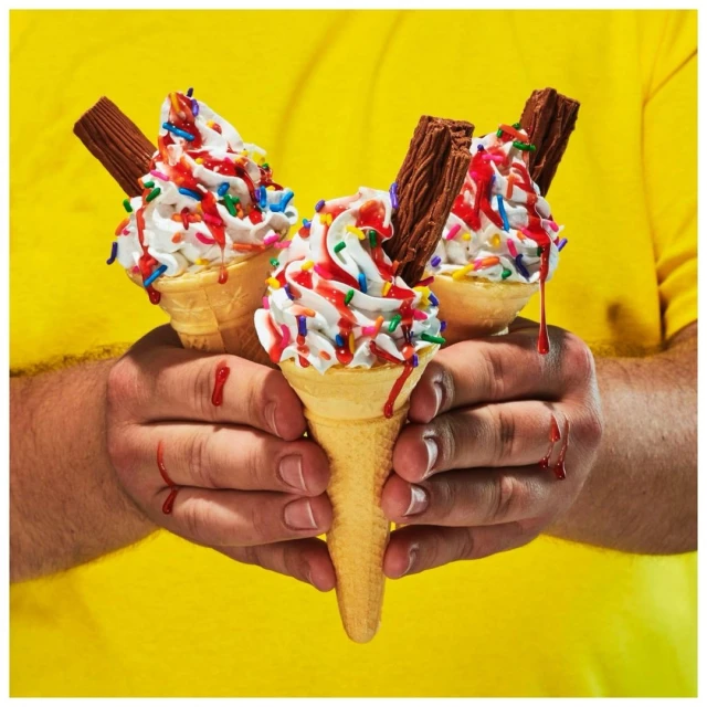 Close-up studio shot of three ice cream cones with whipped cream, sprinkles, sauce, and chocolate sticks held in a person's hands against a yellow backdrop.
