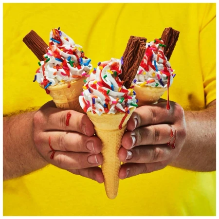 Close-up studio shot of three ice cream cones with whipped cream, sprinkles, sauce, and chocolate sticks held in a person's hands against a yellow backdrop.