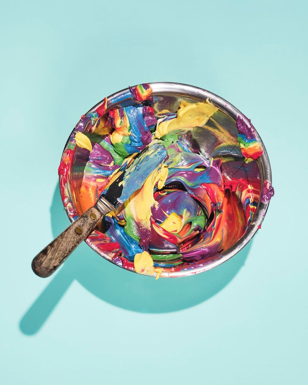 Overhead shot of a metal bowl filled with vibrant, multicolored swirls of paint or frosting, accompanied by a spatula on a light blue background.