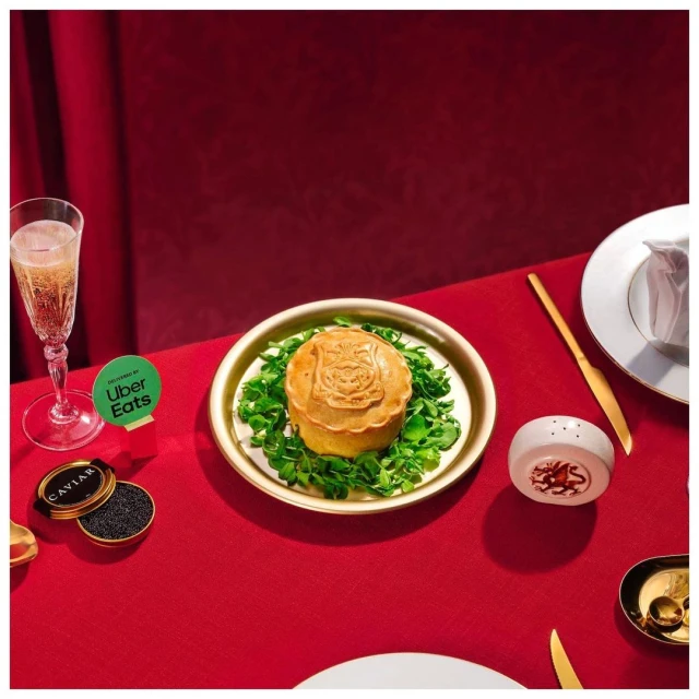Overhead shot of savory meat pie with champagne, caviar, and gold flatware on a red tablecloth, suggesting a luxury food delivery experience.