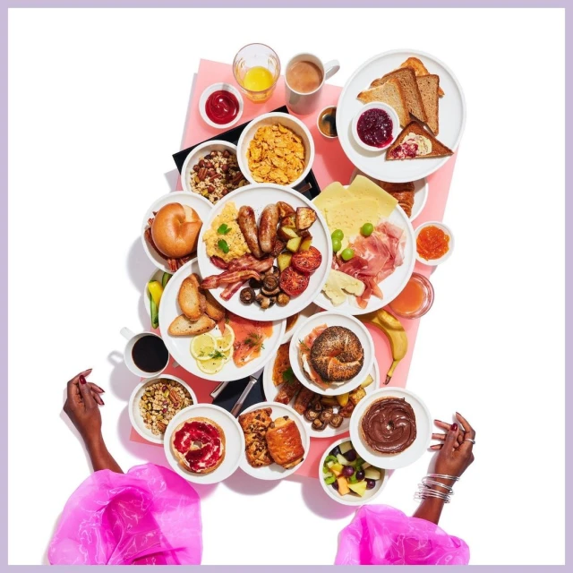 Overhead shot of a woman's hands displaying an array of breakfast foods, including pastries, bagels, fruit, eggs, and breakfast meats.