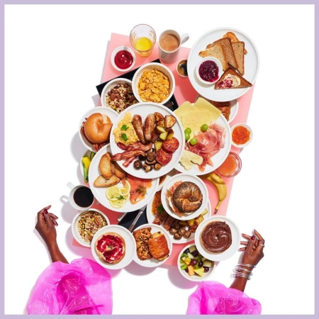 Overhead shot of a woman's hands displaying an array of breakfast foods, including pastries, bagels, fruit, eggs, and breakfast meats.