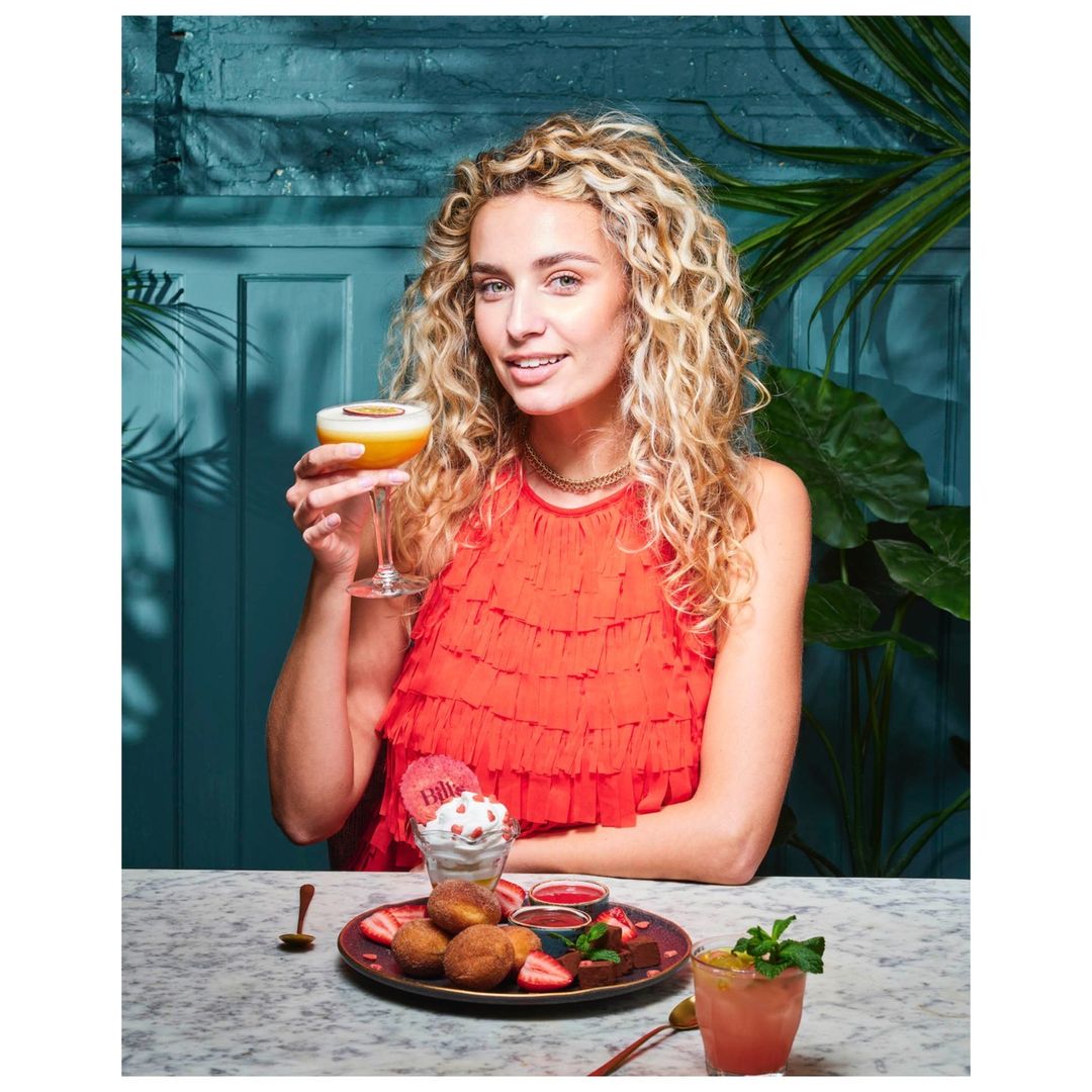 Blonde woman in orange dress holding a cocktail at a marble table with desserts, set against a blue textured wall and tropical plants.