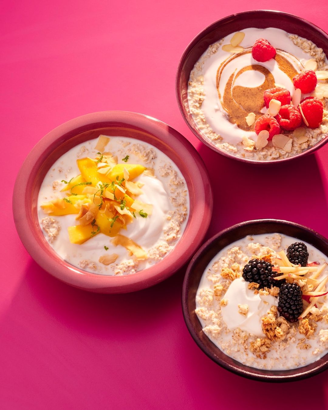 Three bowls of oatmeal with various toppings: mango, coconut, raspberries, almonds, blackberries, apples, and granola on a pink background.