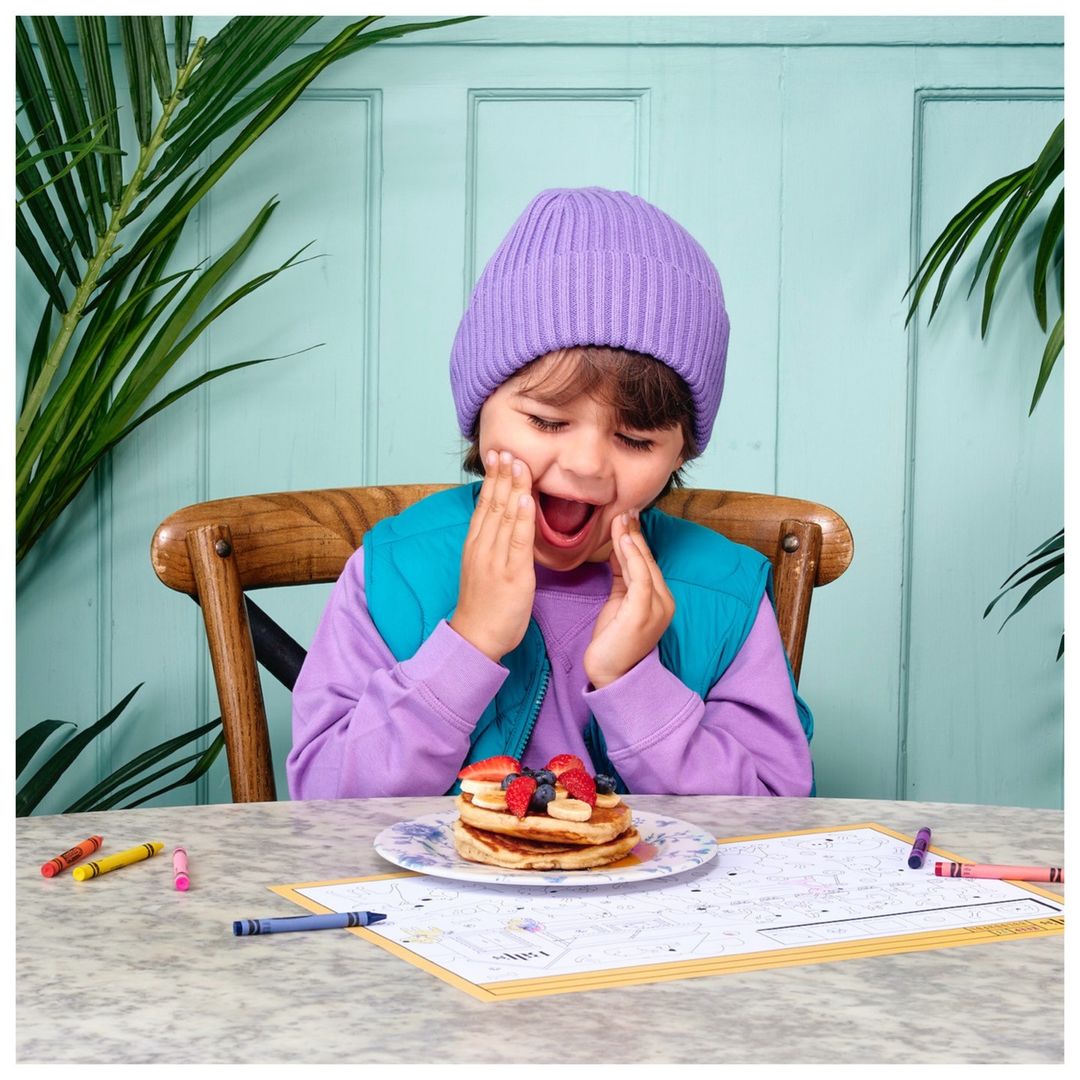Enthusiastic child in a purple hat and vest sitting at a table with pancakes, crayons, and a coloring page inside a home.