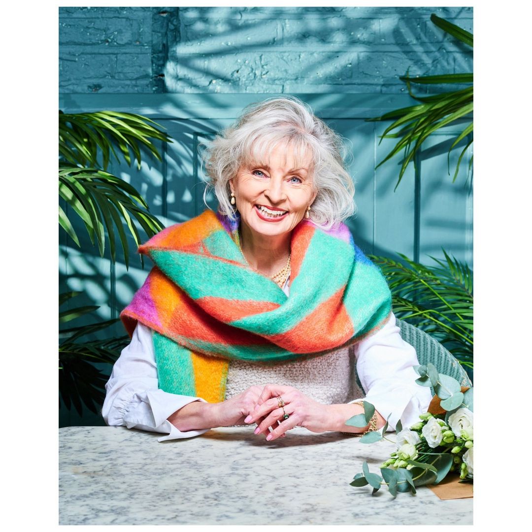 Elegant senior woman with gray hair smiles, wearing a colorful shawl indoors at a table with white roses and green foliage.