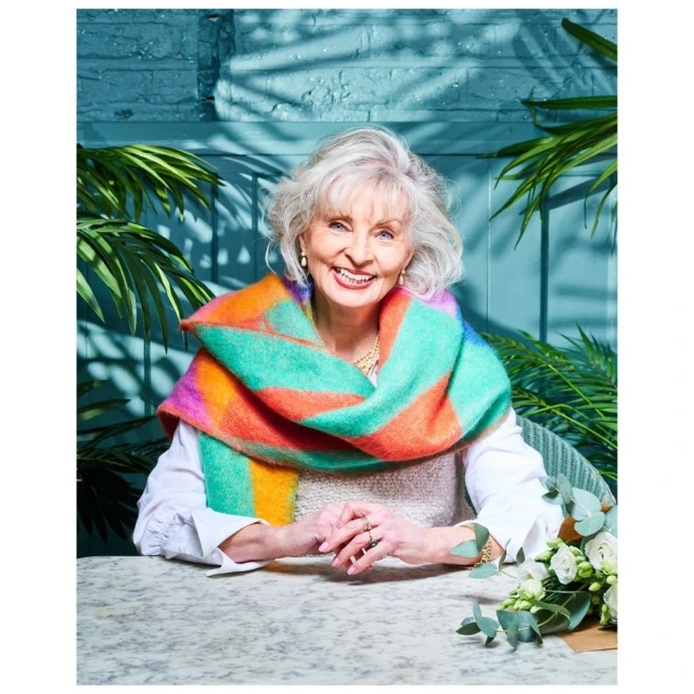 Elegant senior woman with gray hair smiles, wearing a colorful shawl indoors at a table with white roses and green foliage.