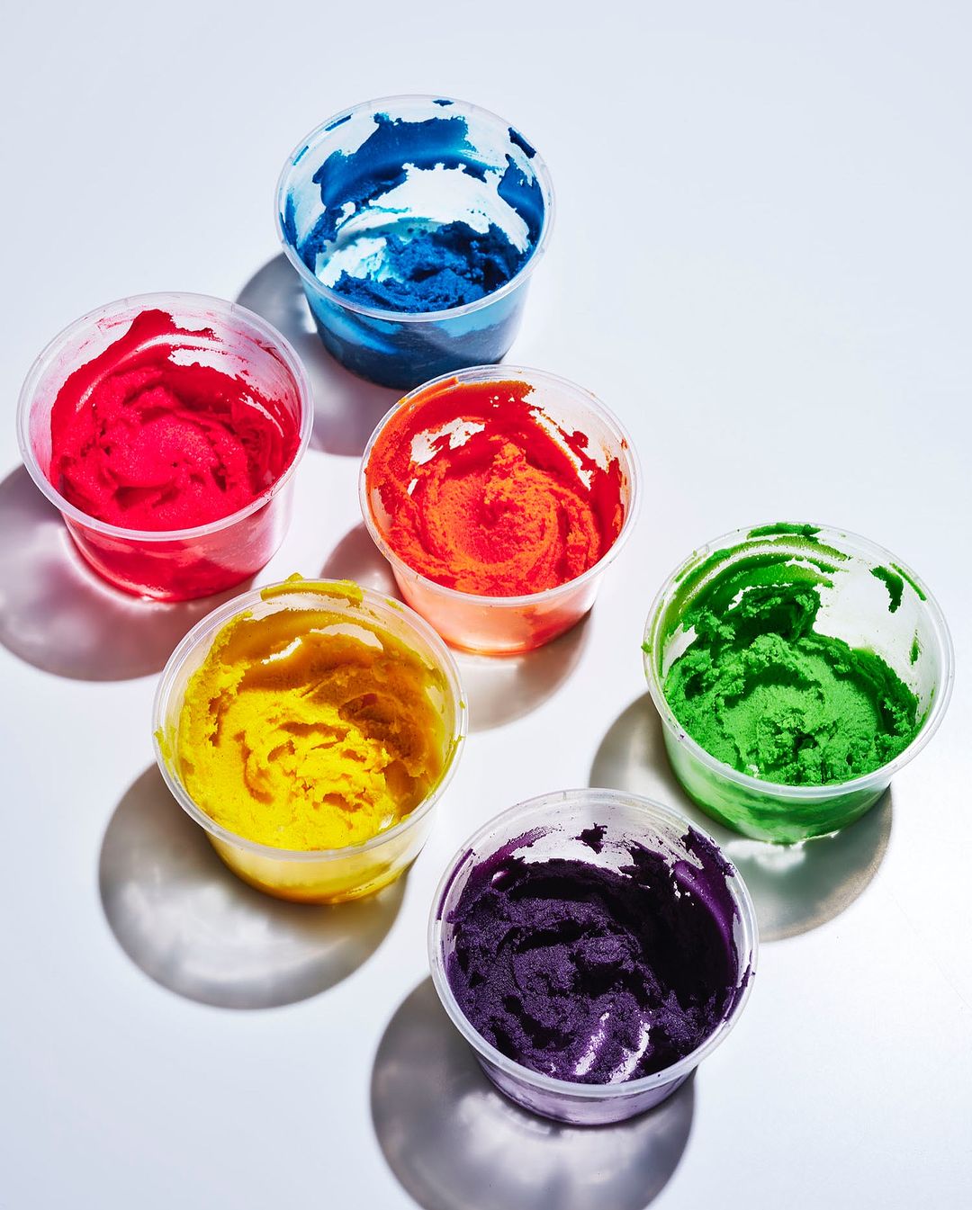 Overhead view of clear plastic containers filled with brightly colored rainbow dye or pigment against a bright white background.