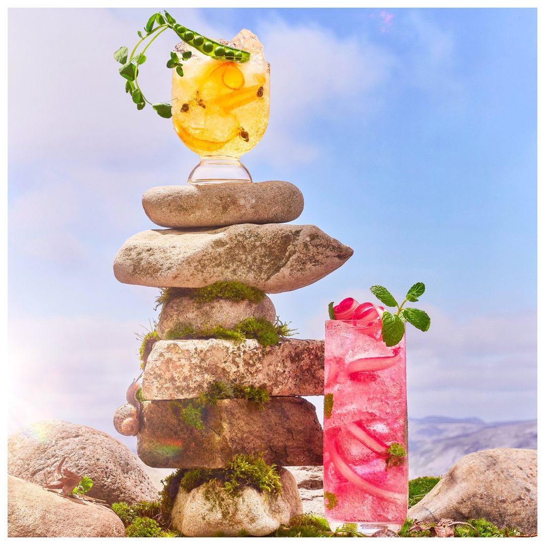 Two vibrant cocktails are balanced on a mossy rock cairn under a blue sky, evoking fresh, natural, and refreshing beverage photography.